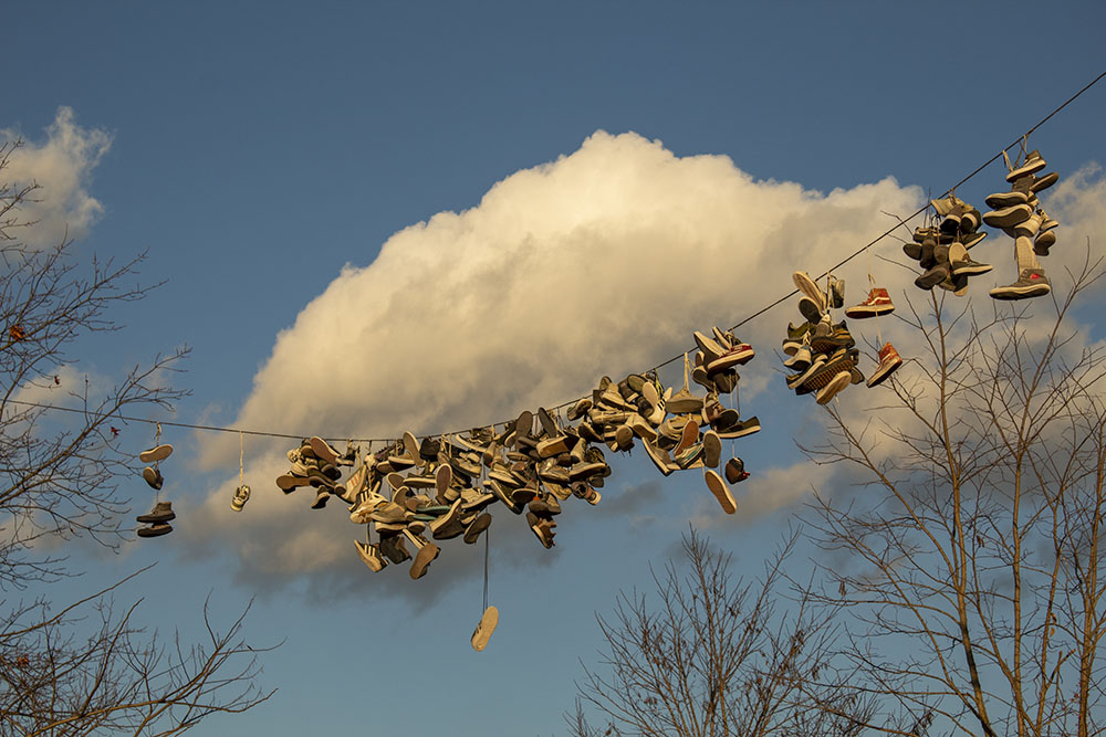 The line of a million miles. A photo of a line of dozens a shoes hanging off of it. There is a big cloud in the background and some trees with no leaves.